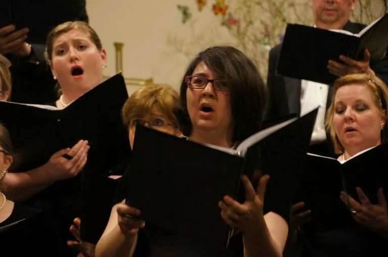 Jamie holds up her music folder and sings with an intense look on her face, surrounded by other choir members on stage.