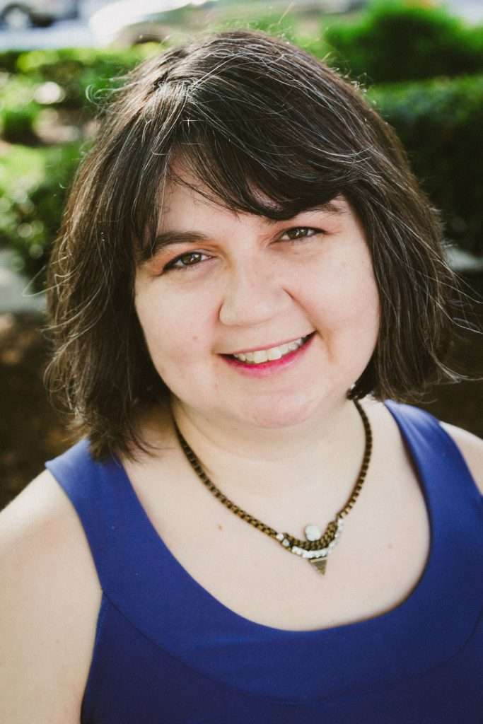 Jamie, a white woman with chin-length dark hair and bags, looks at the camera and smiles. She's wearing a sleeveless indigo dress and a statement necklace.